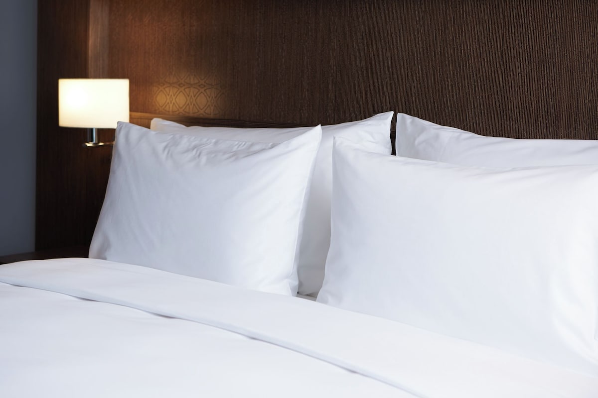 A neatly made bed with white linens, a wooden headboard, and a wall-mounted lamp.