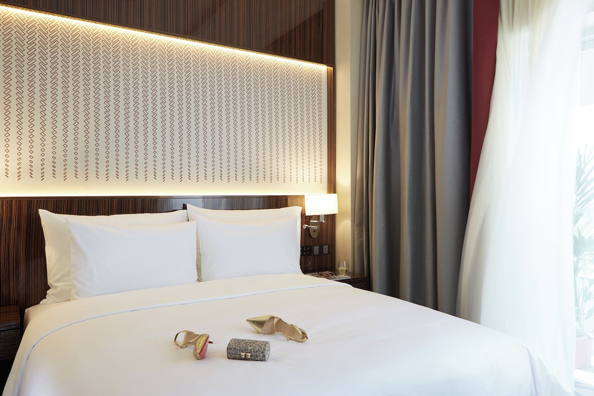 A neatly made bed with white linens, a headboard with a geometric pattern, and a nightstand with a lamp and a glass of water.