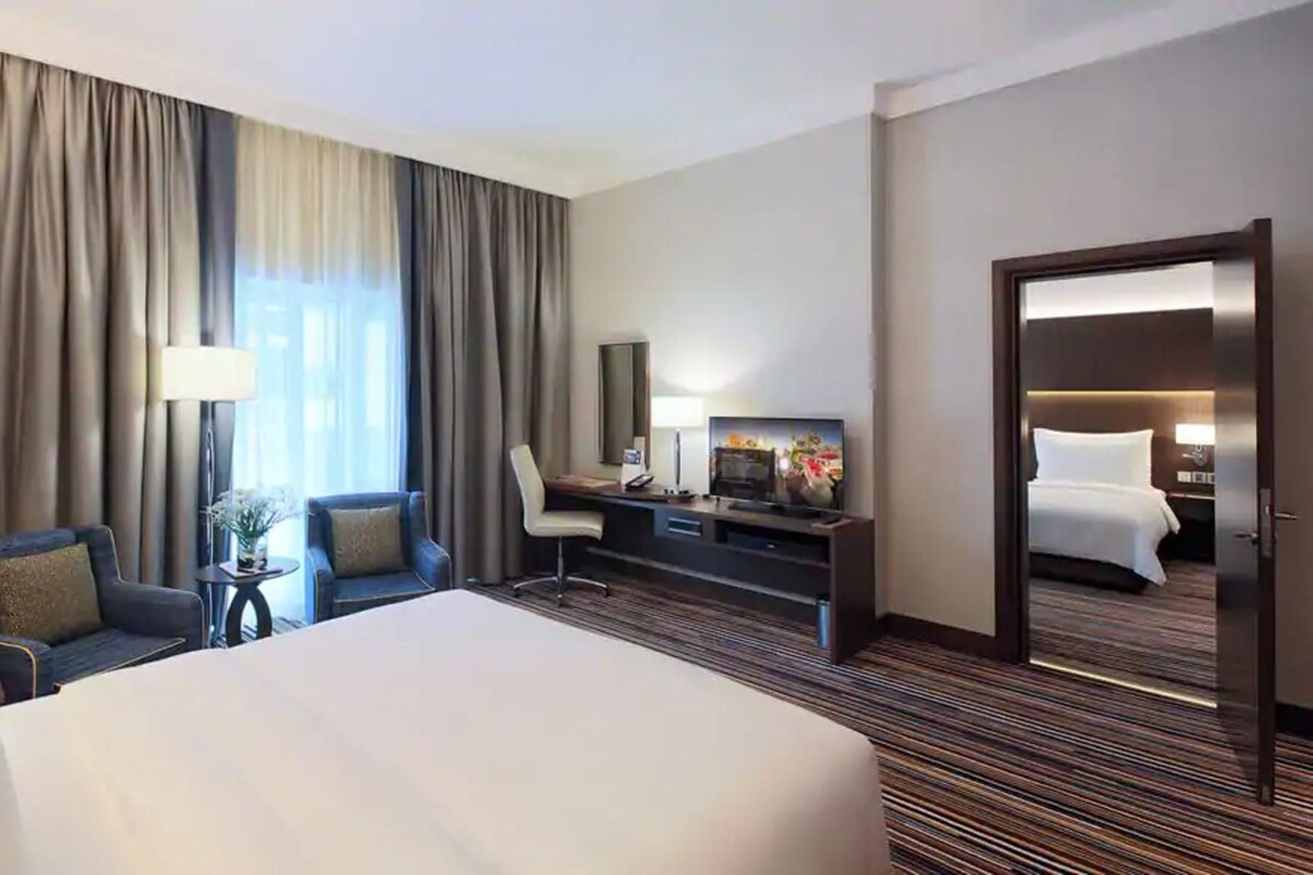 A hotel room with a large bed, a desk with a TV, a chair, a lamp, and a door leading to another room.