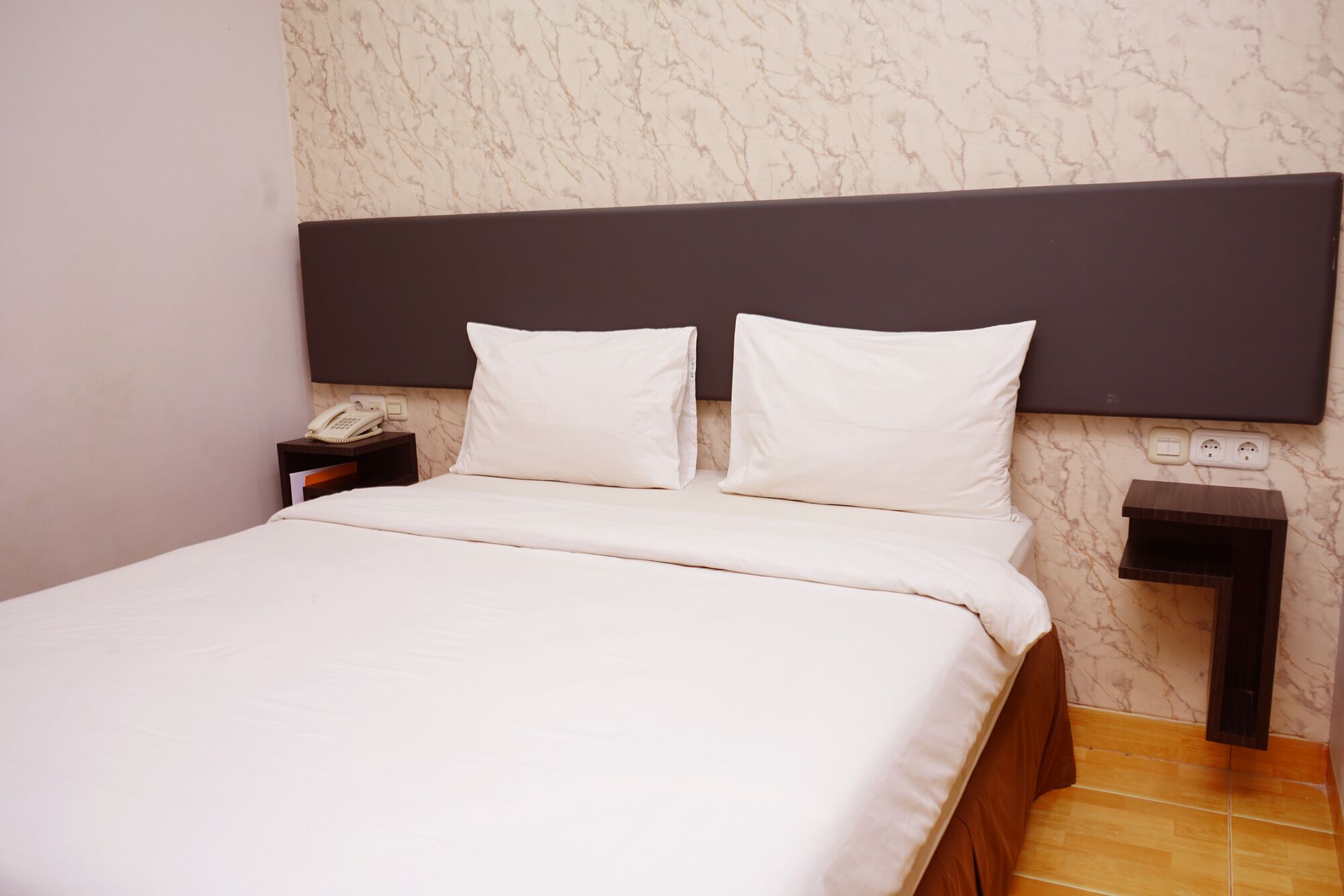 A neatly made bed with white linens and pillows, a bedside telephone, and a nightstand with a power outlet.