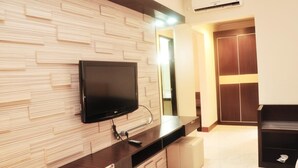 A modern living room with a flat-screen TV mounted on a wooden wall, a dark wooden console table, a beige ottoman, and a wardrobe with sliding doors. - Hotel 88 Diponegoro (Jember)