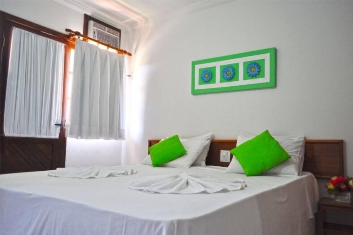 Une chambre d’hôtel avec un lit, des rideaux blancs, un élément de décoration murale vert et une table de chevet sur laquelle se trouve un vase de fleurs.