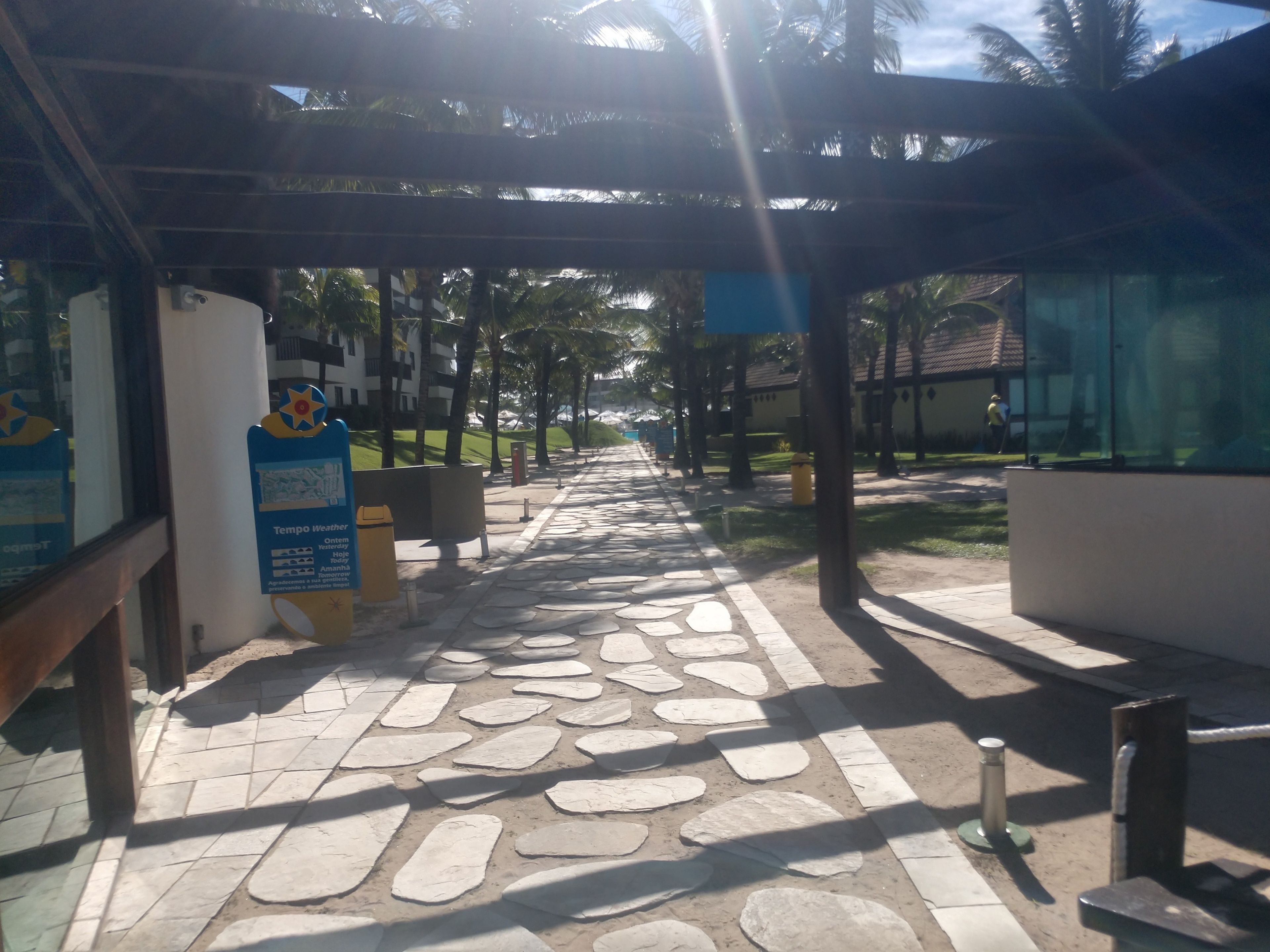 A covered walkway with stone tiles, palm trees, and a weather sign.