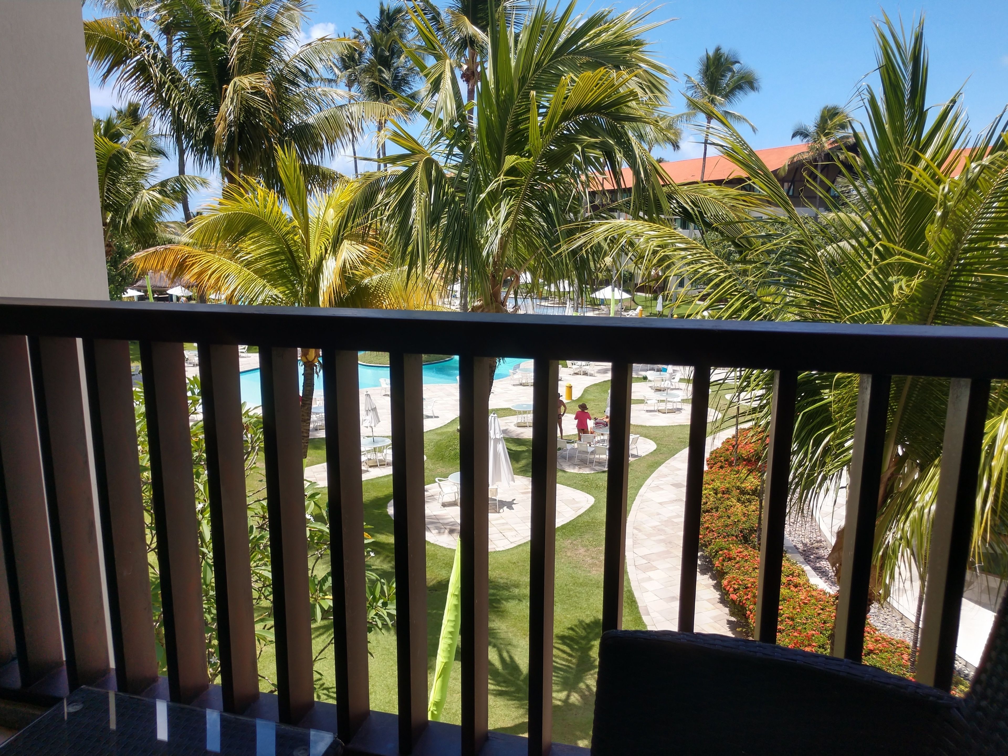 Ein Balkon mit Blick auf einen Poolbereich, Palmen und ein Gebäude mit rotem Dach.