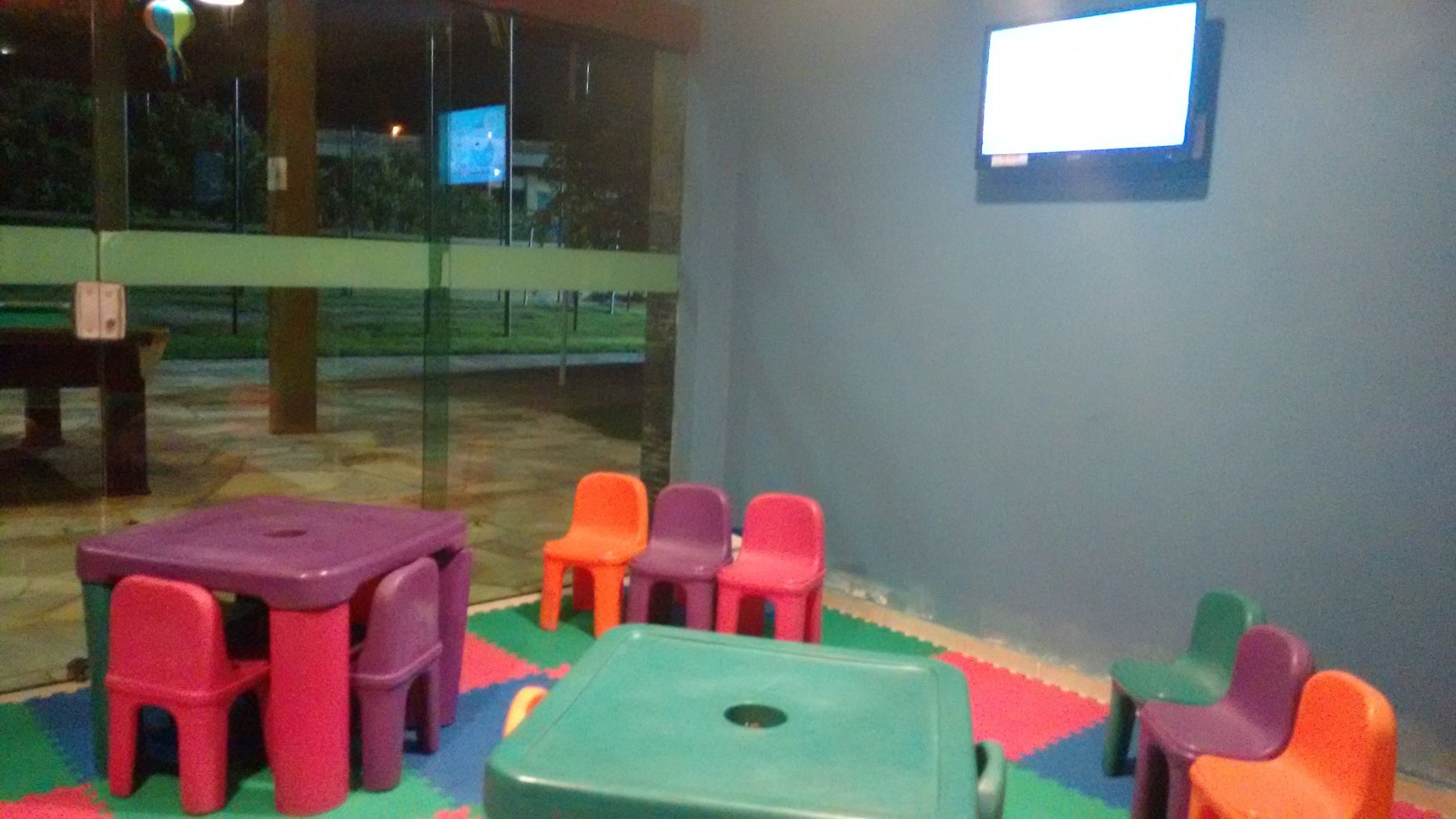A children's play area with colorful plastic chairs and tables, a television on the wall, and a glass door leading outside.