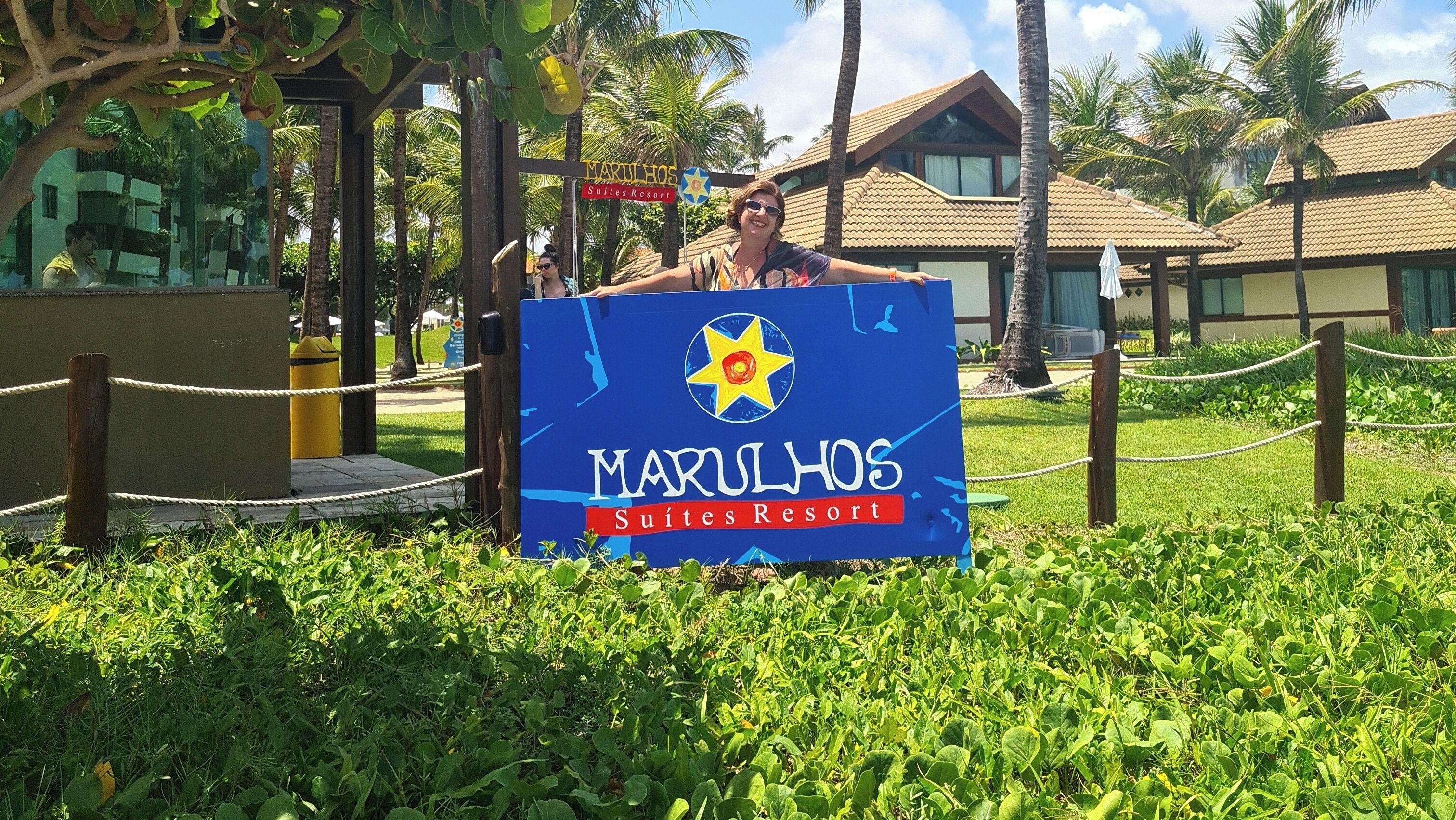 A sign for Marulhos Suites Resort with a star emblem, surrounded by greenery and palm trees.