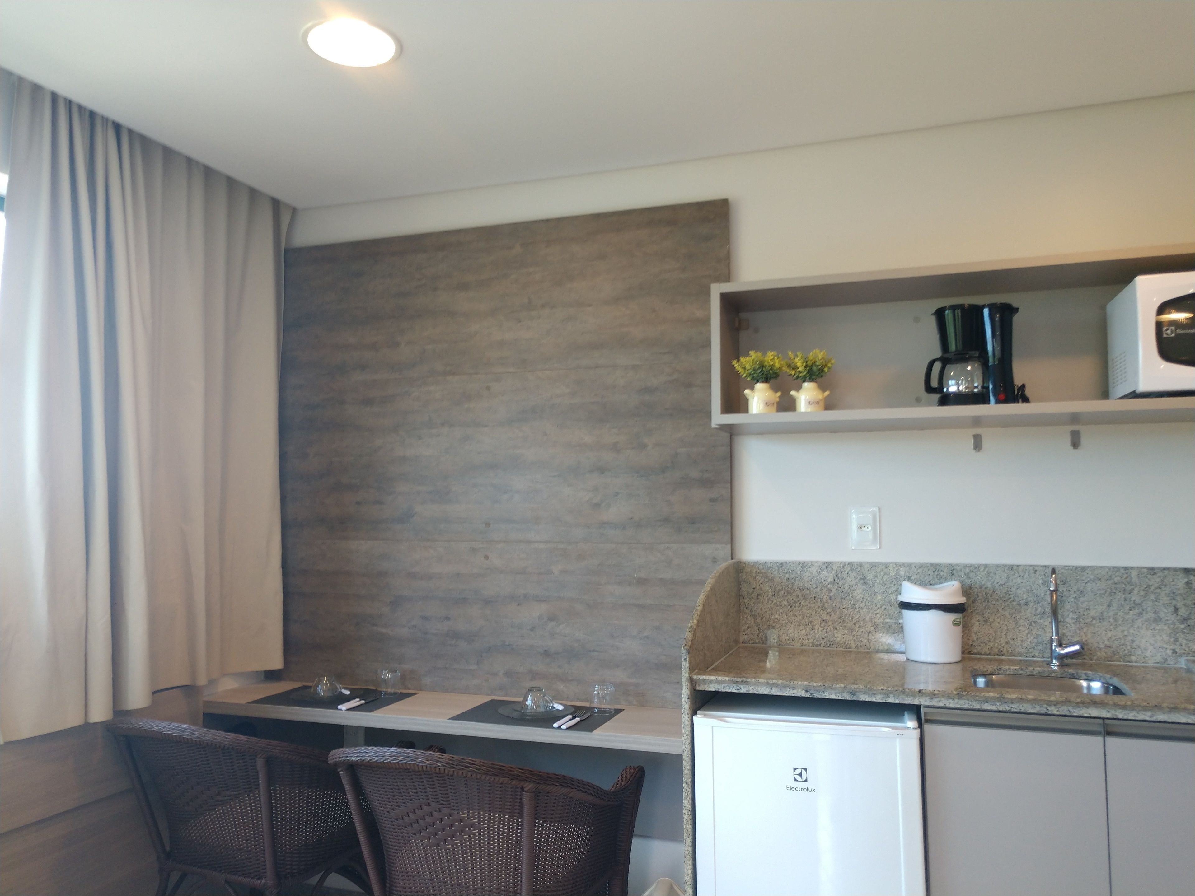 A compact kitchen with a built-in refrigerator, a microwave, and a coffee maker on a shelf. There is a dining table with chairs and a small potted plant on the shelf.