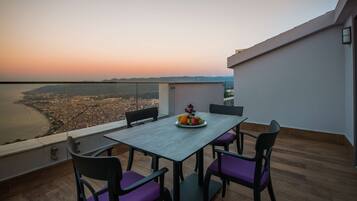 Un balcón con una mesa lista para dos personas, que incluye un plato de fruta y vistas a una ciudad costera al atardecer.