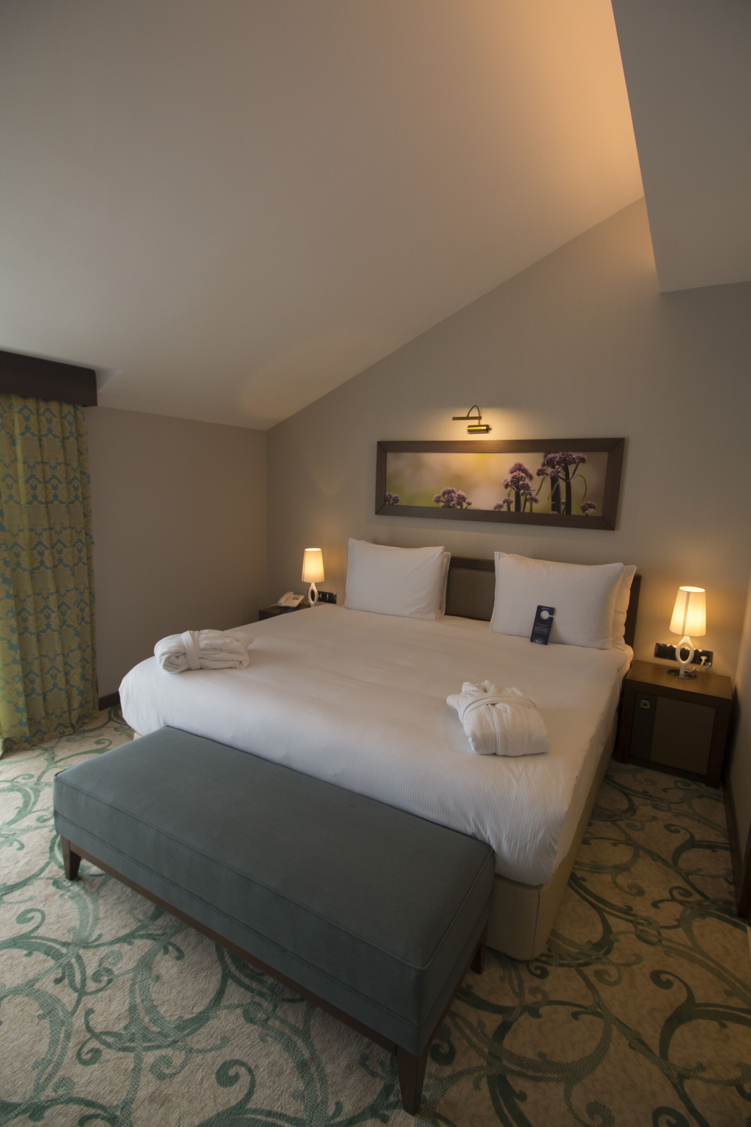 A hotel room with a large bed, two bedside tables with lamps, a bench at the foot of the bed, a painting above the bed, and a patterned carpet.