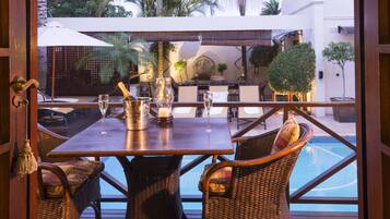 An outdoor dining area with a wooden table and wicker chairs, a pool, and a view of the surrounding landscape.