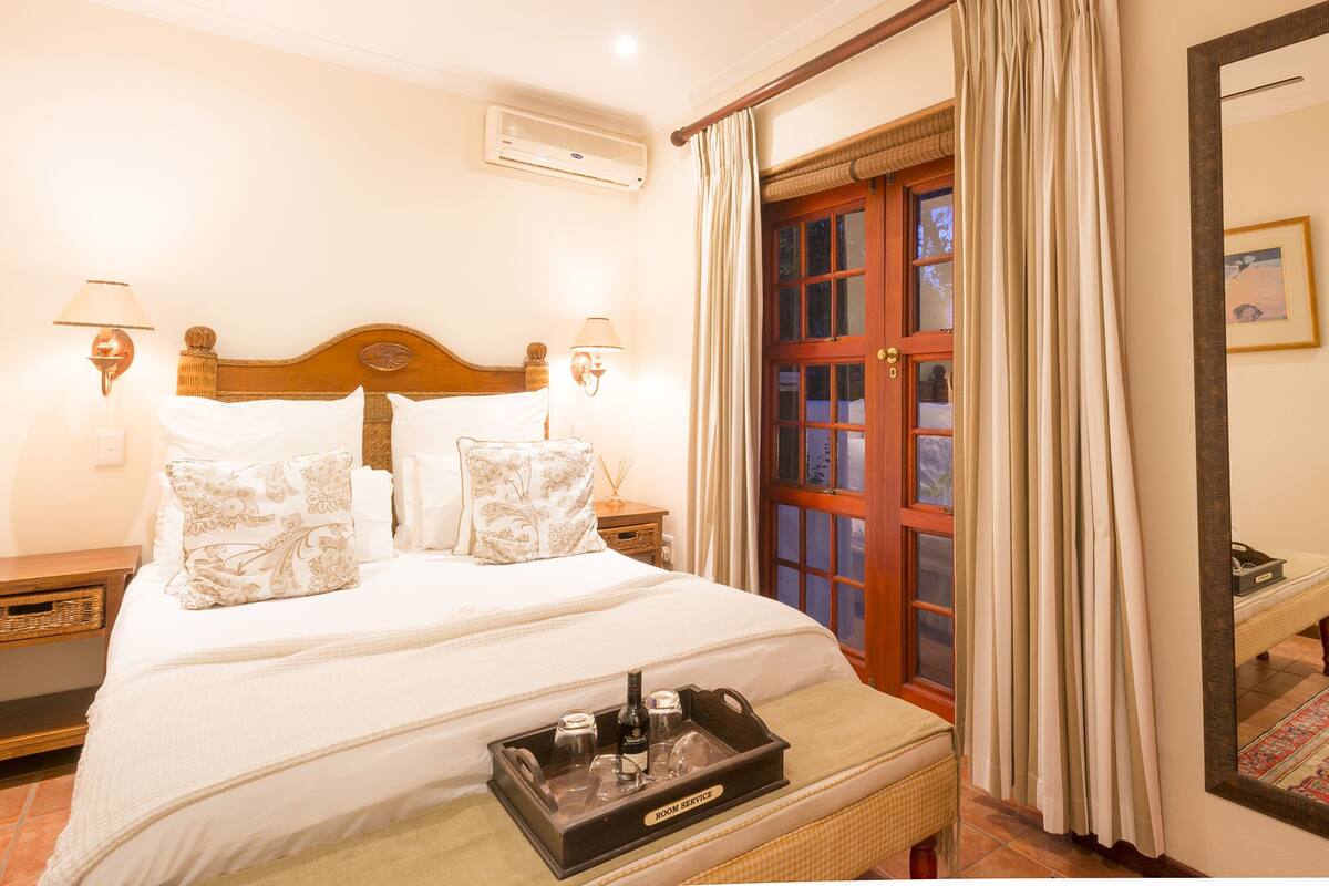 Una habitación de hotel con una cama, una bandeja con una botella y vasos, un cabecero de madera, dos lámparas de pared, una ventana con cortinas y un espejo.