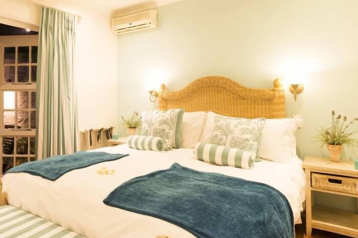 Un dormitorio con cama, almohadas, mesita de noche y ventana con cortinas.
