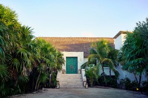 A building with a green door, surrounded by palm trees and a thatched roof structure in the background.