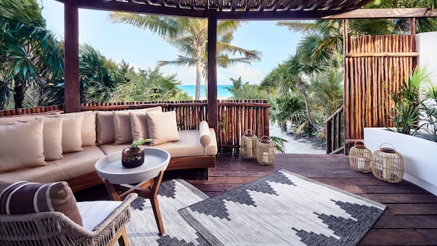 A covered outdoor seating area with a sofa, chairs, and a table, overlooking a beach with palm trees and a wooden fence. - Casa Chablé Sian Ka'an Reserve - South of Tulum (Punta Allen)