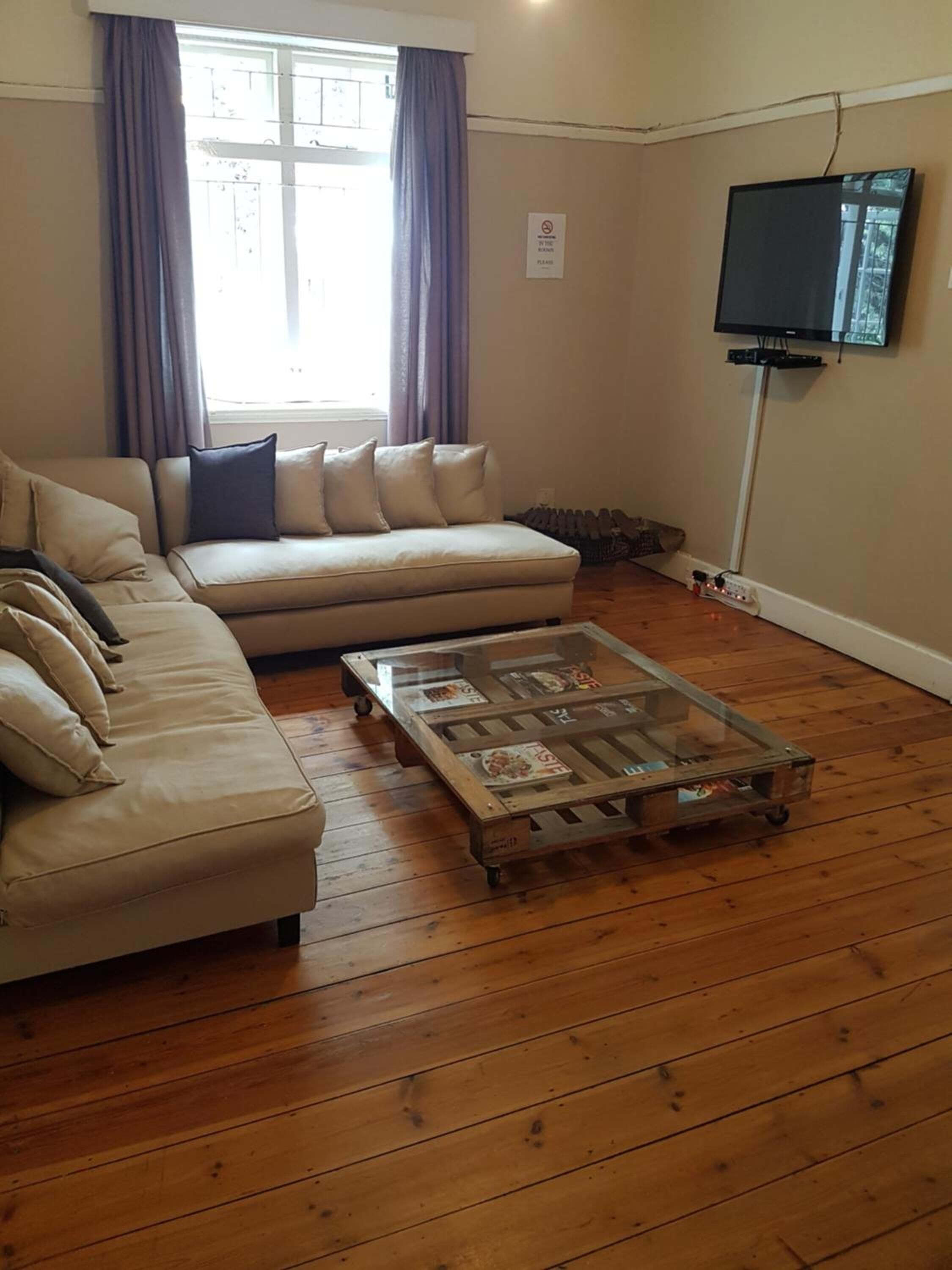 A living room with a sofa, a glass coffee table, wooden flooring, a television, and a window with curtains.