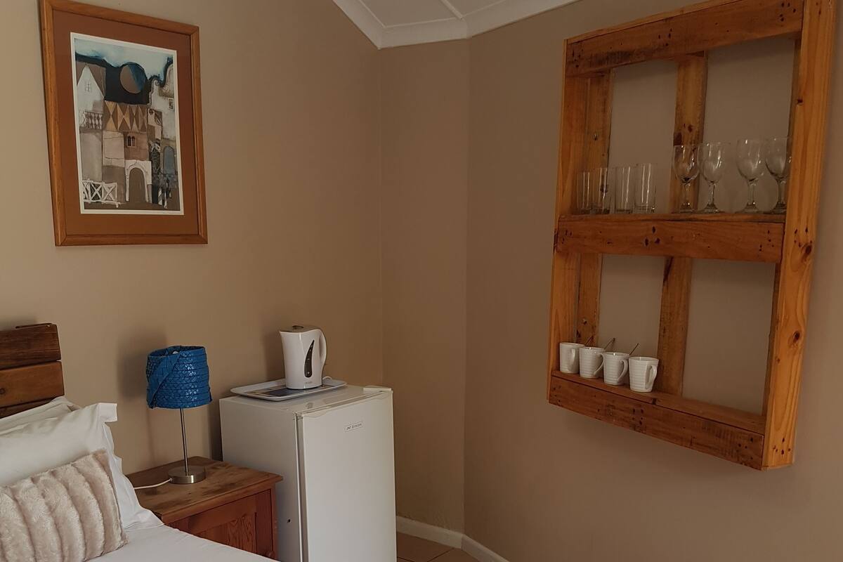 A bedroom with a bed, a desk with a lamp, a refrigerator, a framed picture on the wall, and a wooden shelf with glasses and cups.