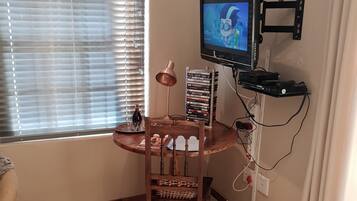 Una habitación con televisión montada en la pared, una silla de madera, una mesita pequeña y una ventana con persianas.