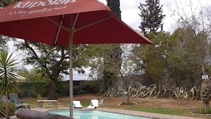 Zona de piscina con un banco de madera y un paraguas rojo, rodeada de árboles y un patio de ladrillo.