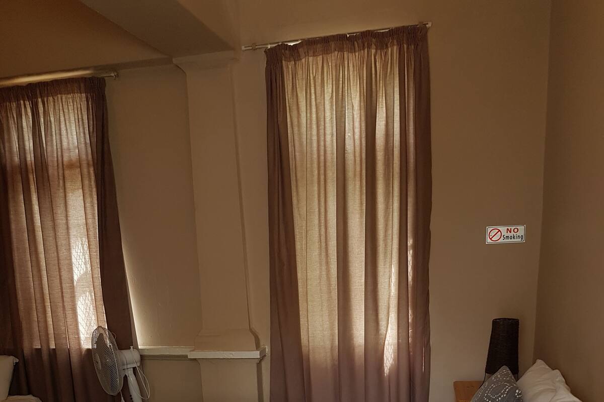 A bedroom with a bed, curtains, a fan, a wooden nightstand, and a 'No Smoking' sign.