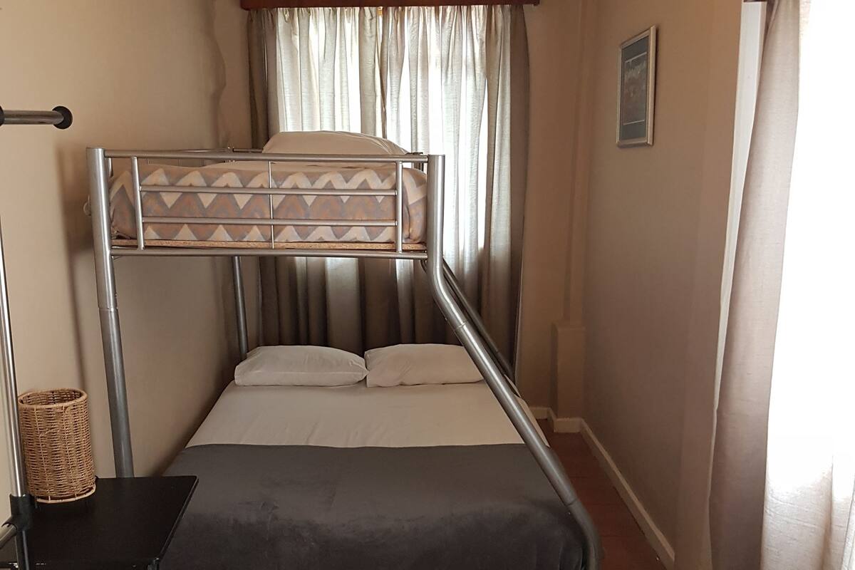 A compact room with a bunk bed, a small desk, a fan, and a window with curtains.