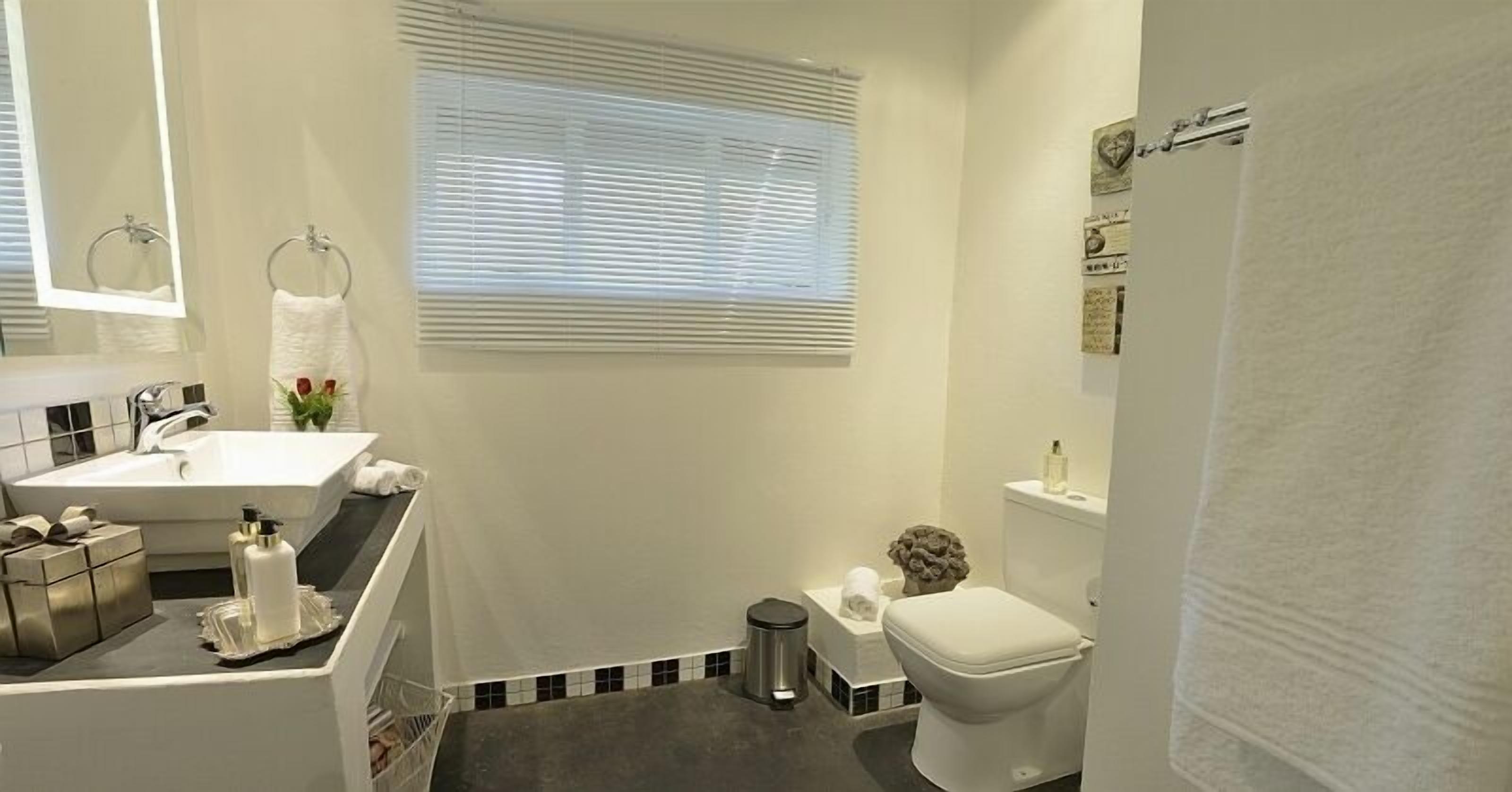 A modern bathroom with a white sink, vanity, and toilet. There is a window with blinds, a small trash bin, and a towel rack.