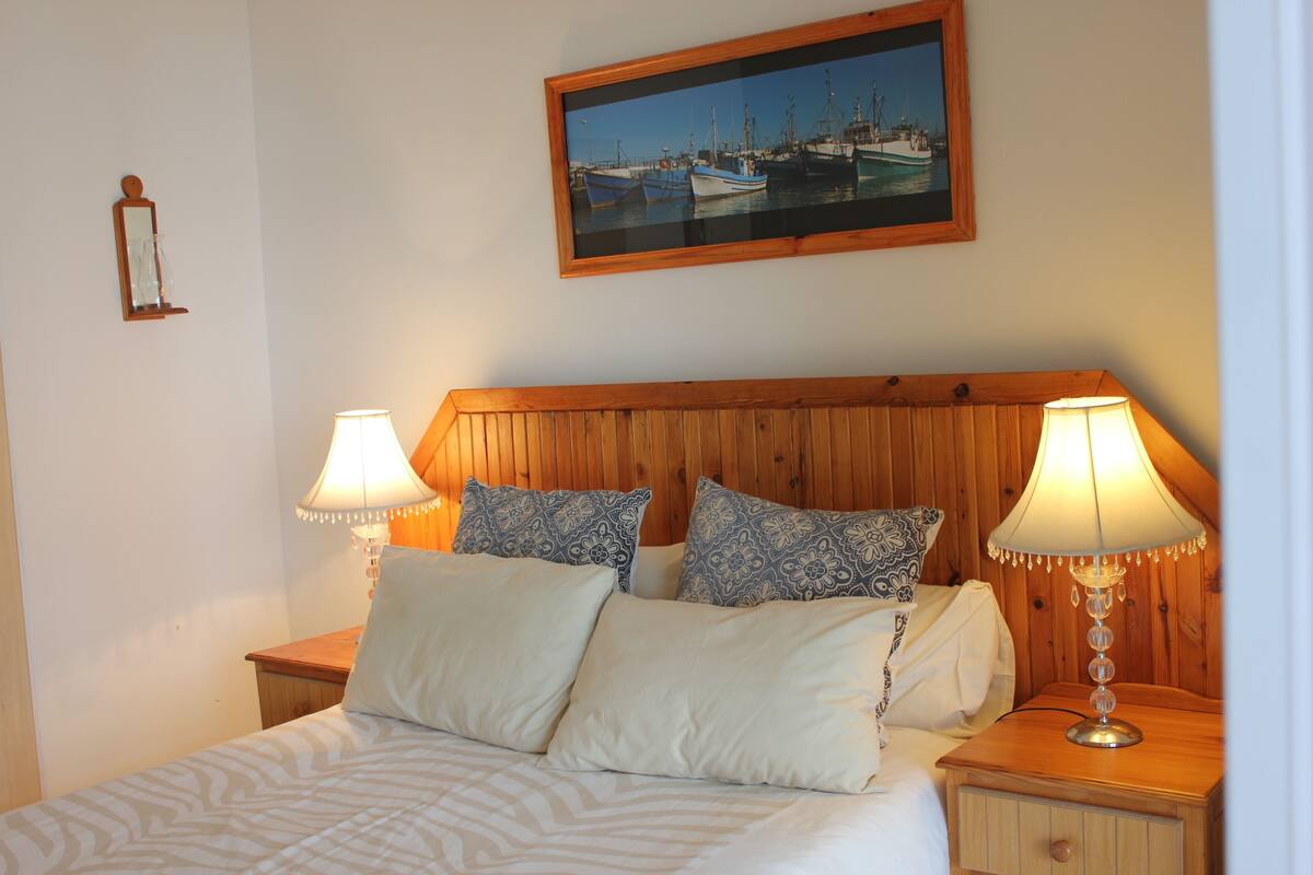 Una cama bien hecha con ropa de cama estampada, dos lámparas de noche y un cuadro con barcos en la pared.