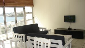 Une pièce avec une table et des chaises blanches, un canapé noir, un téléviseur posé sur un meuble noir, et une vue sur la mer par de grandes fenêtres.