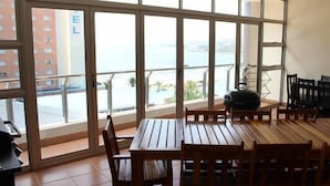 Un coin repas avec une table et des chaises en bois, de grandes fenêtres donnant sur la mer et les bâtiments, et une bouilloire posée sur la table.