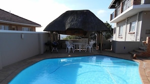 A swimming pool with a thatched roof structure, surrounded by white chairs and tables, and adjacent to a building with a balcony. - Hamelin Guest House (Bluff)
