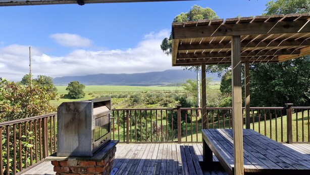 Een houten terras met een barbecue en een pergola, met uitzicht op een groen landschap en verre bergen.
