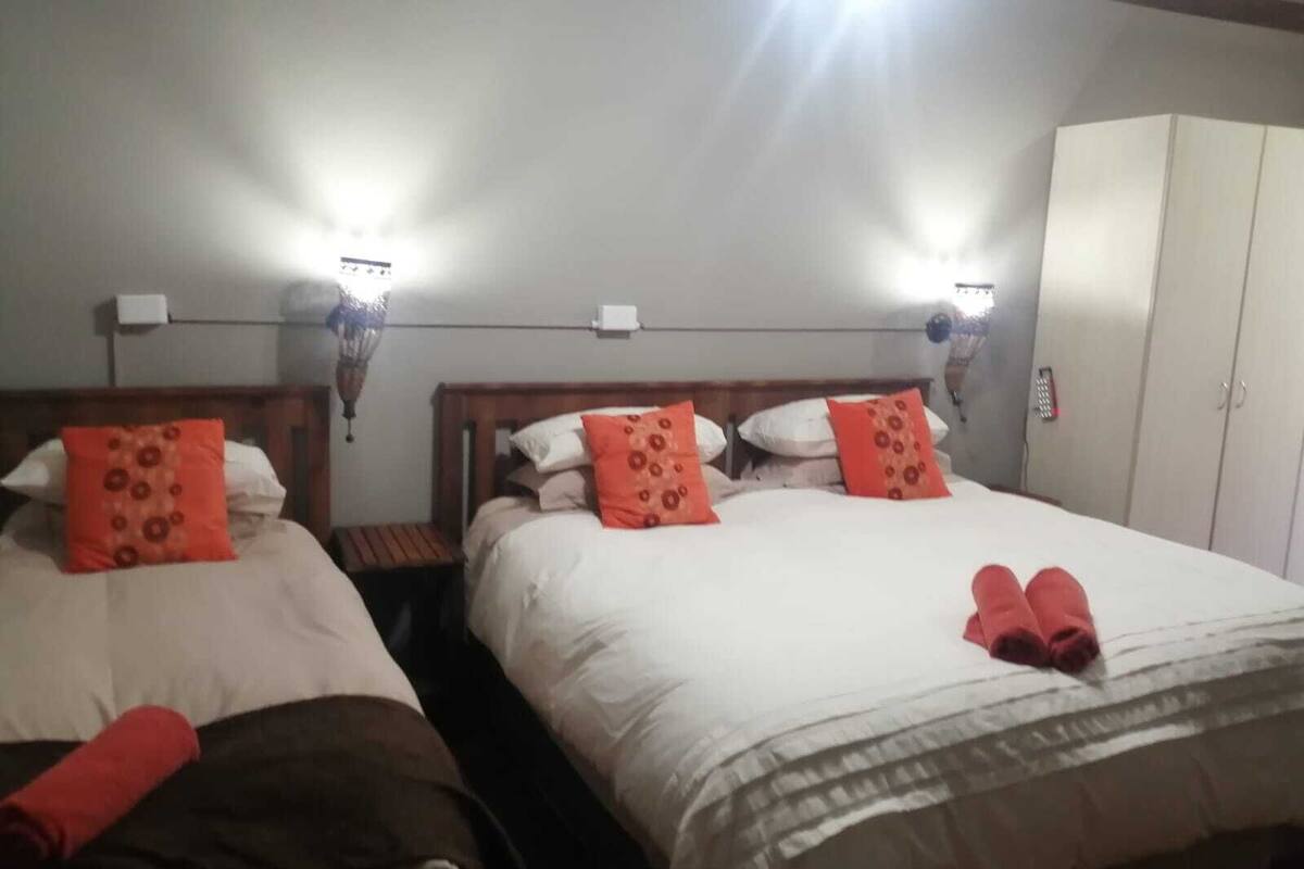 A hotel room with two beds, each with a wooden headboard, white bedding, and orange pillows. There are wall-mounted lights above each bed, and a white wardrobe is visible in the background.