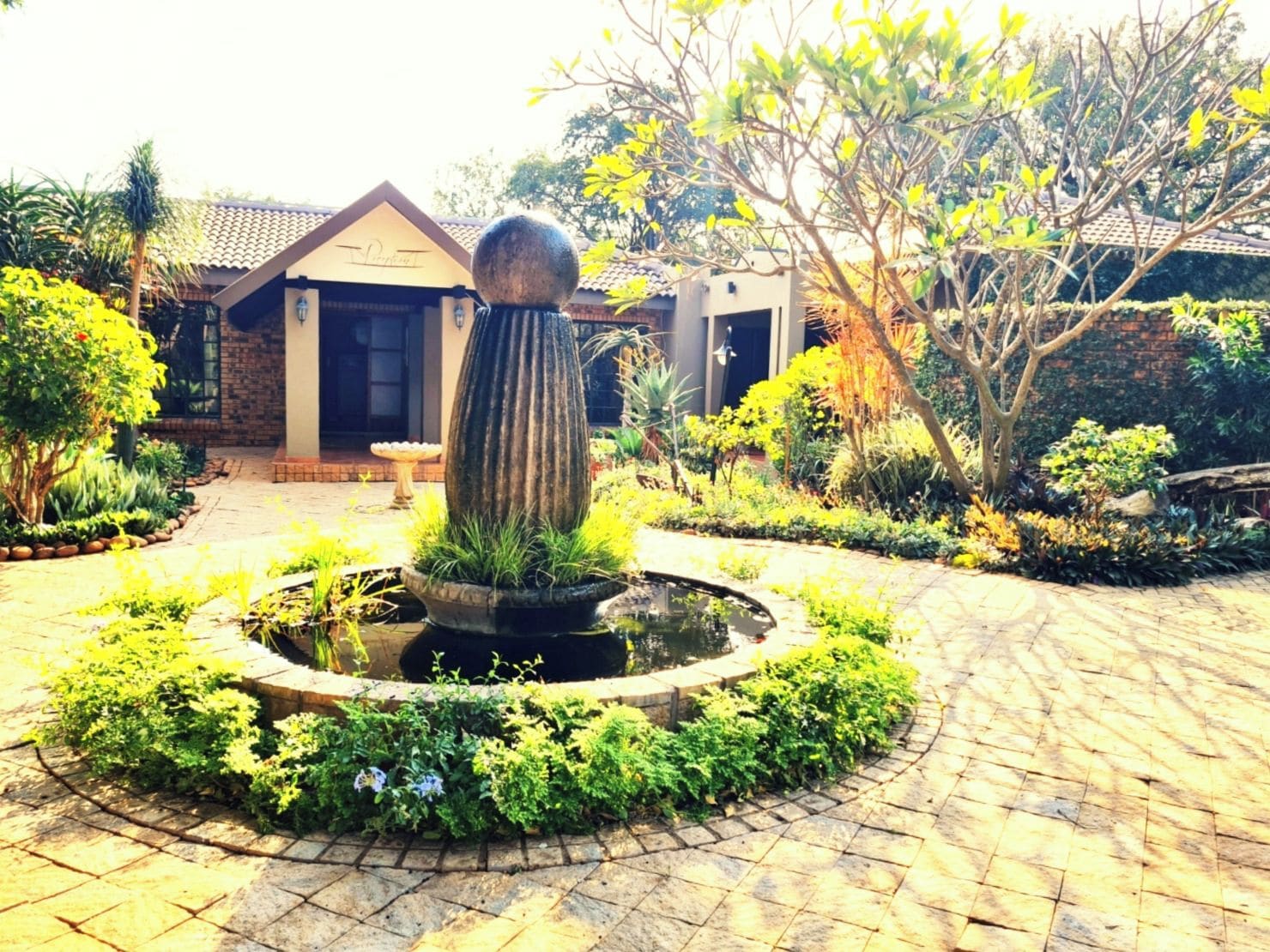 Un patio con una fuente central, rodeado de abundante vegetación y un edificio con techo de tejas.