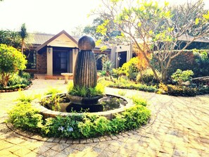A courtyard with a central water feature, surrounded by lush greenery and a building with a tiled roof. - Barnstormers Rest (Nkomazi)