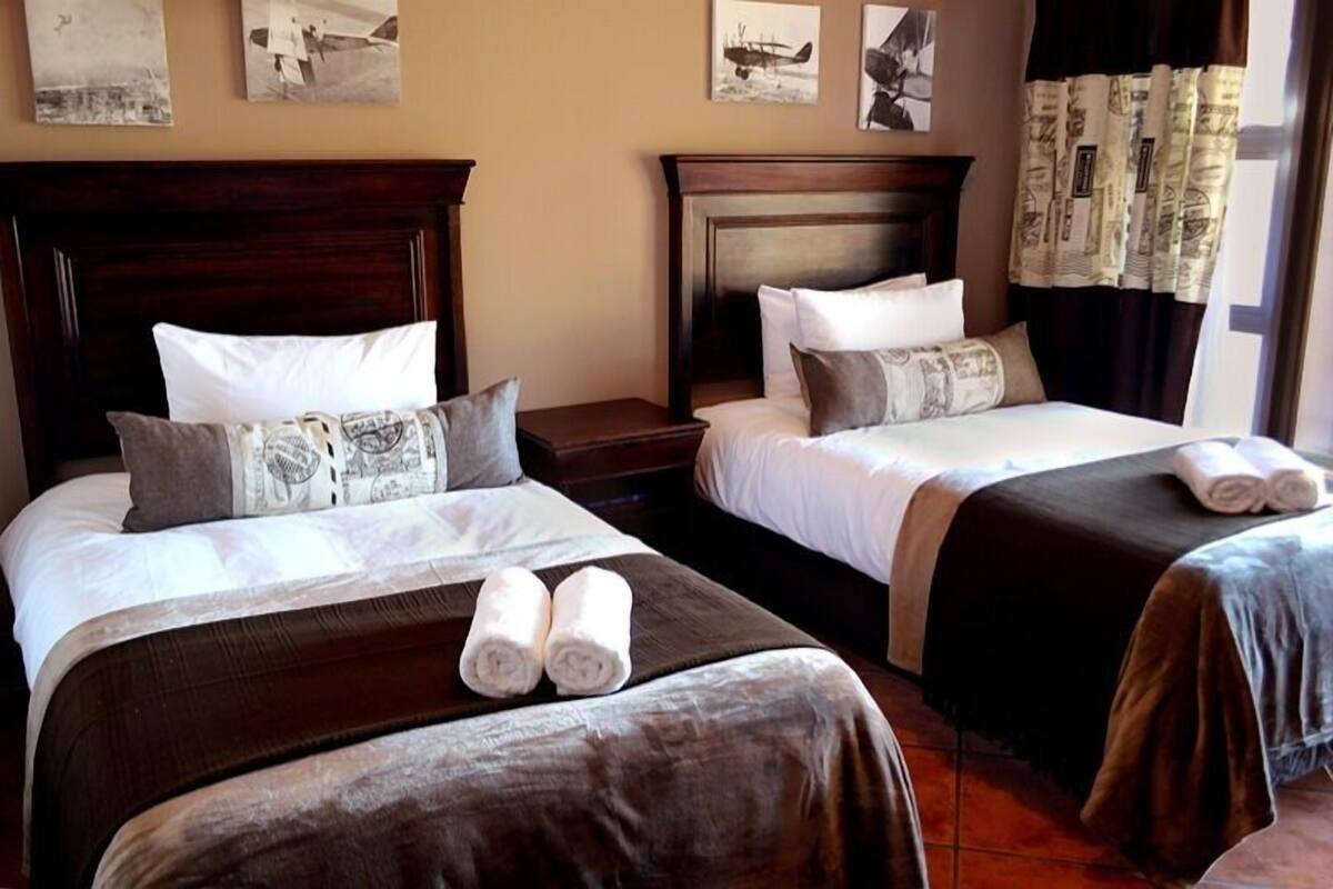 A hotel room with two beds, each with a wooden headboard, white bedding, and a brown bedspread. There are two rolled towels on the beds.