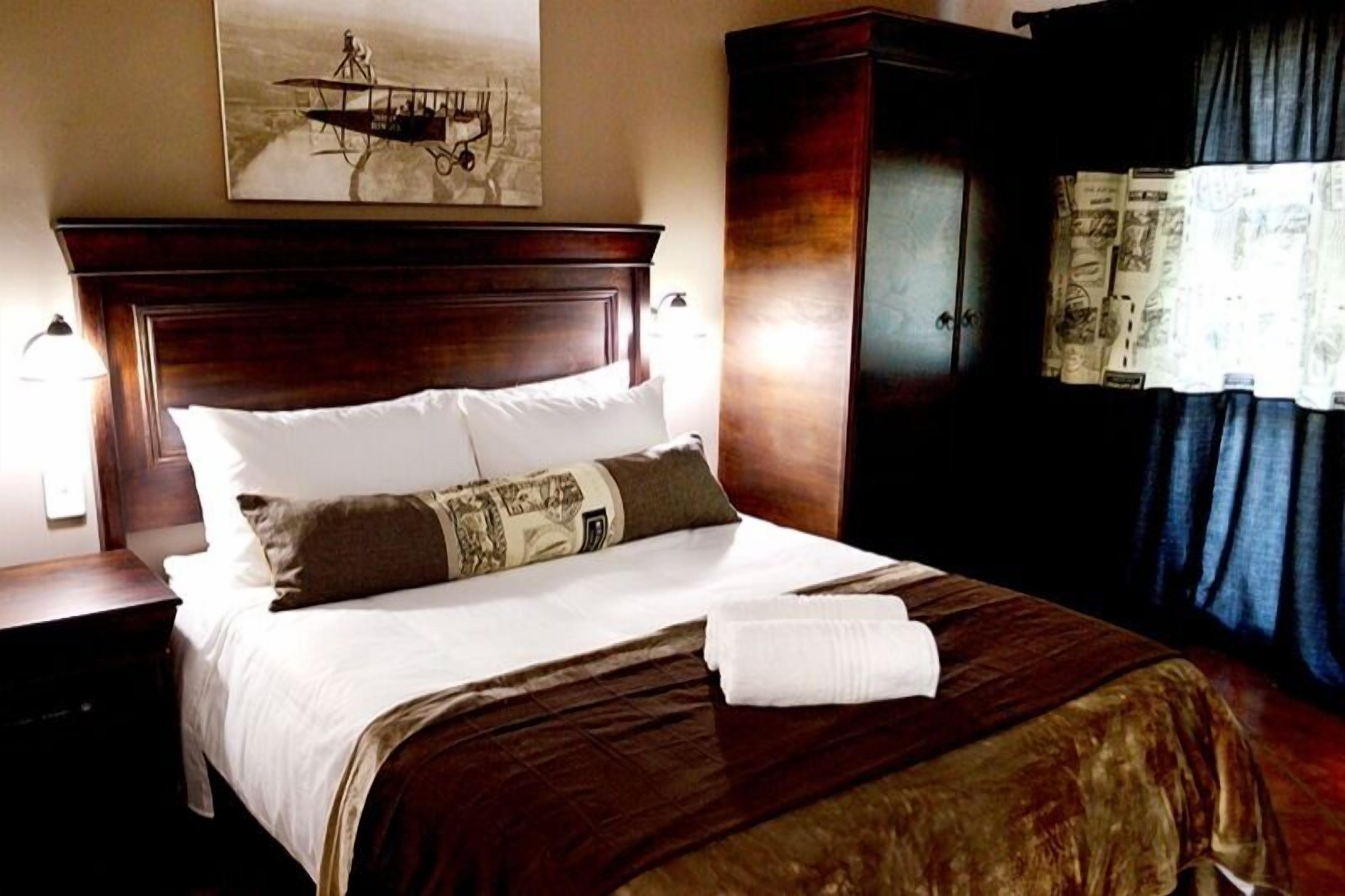 A neatly made bed with a brown headboard, white pillows, and a decorative pillow. A white towel is placed at the foot of the bed.