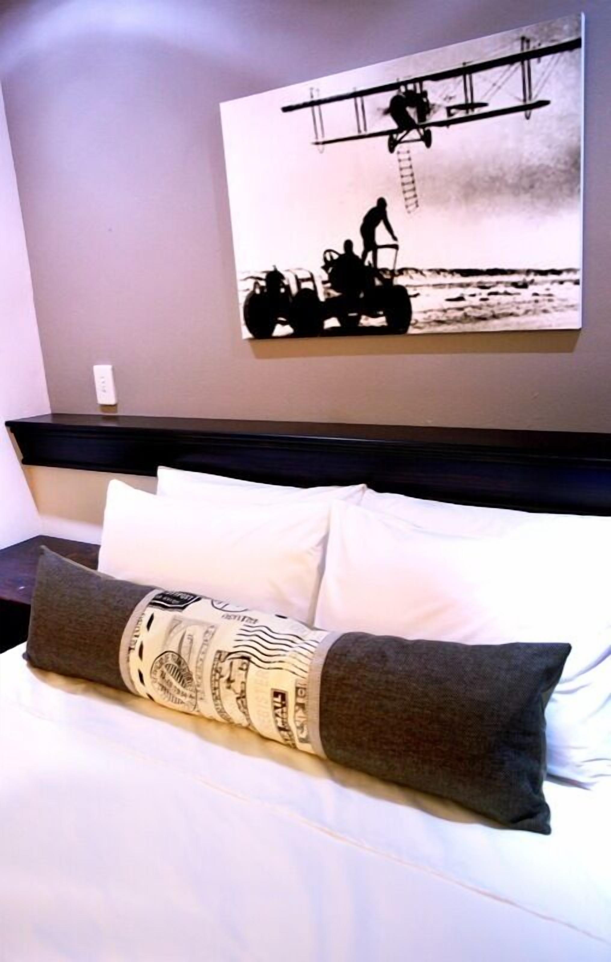 Ein Hotelzimmer mit einem Bett, das ein Kopfkissen mit einem Dollar-Motiv, eine Kopfteil und ein an der Wand befestigtes Kunstwerk zeigt, das eine Person auf einer Leiter darstellt.