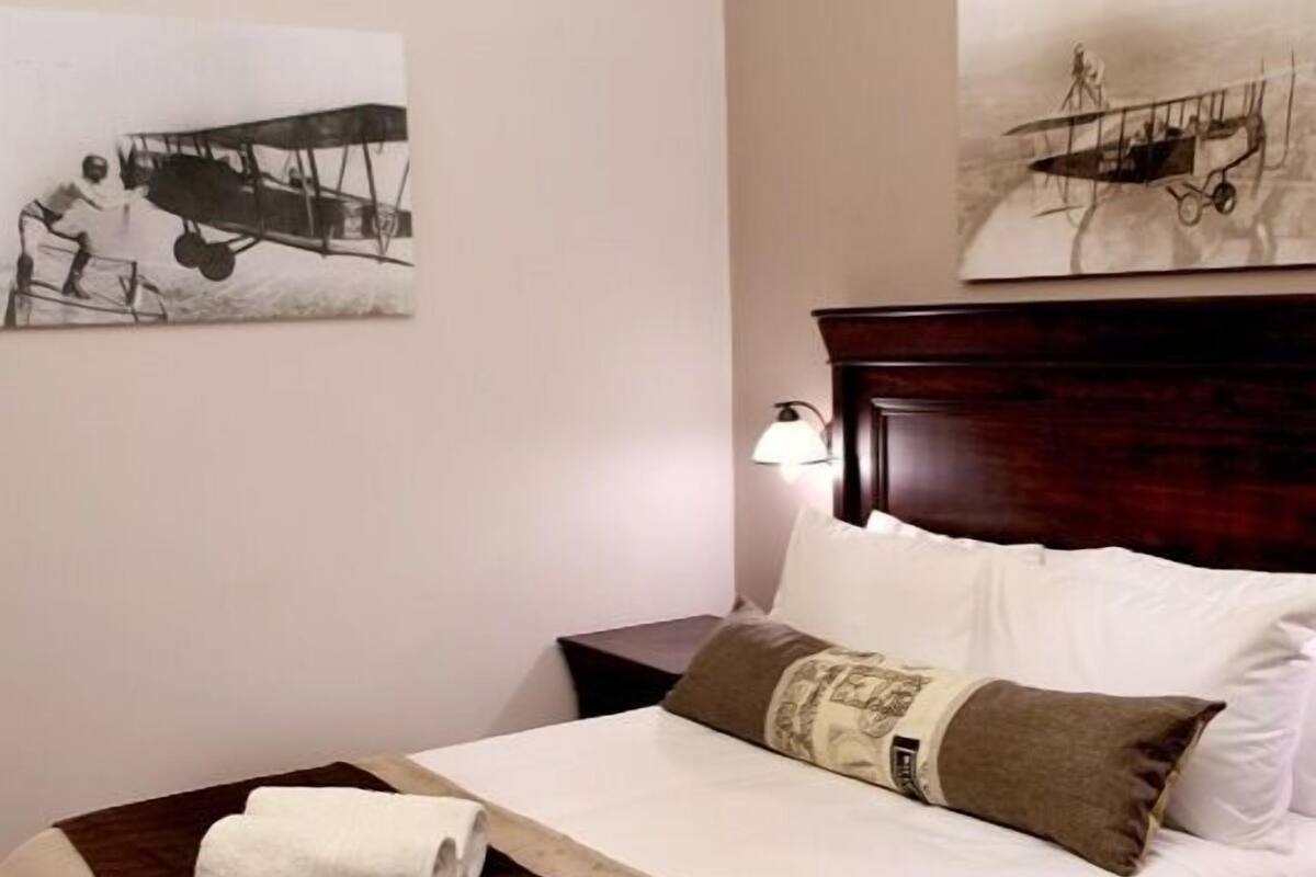 A neatly made bed with a white and brown bedspread, two pillows, and a folded white towel. There are two bedside tables with lamps, a wooden headboard, and two wall-mounted light fixtures.