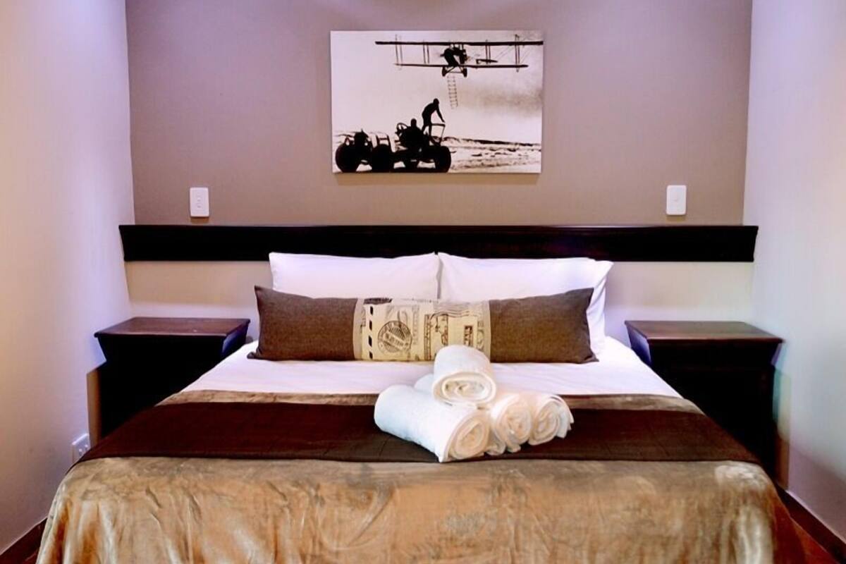 A neatly made bed with a brown bedspread, white pillows, and a decorative pillow with a dollar bill design. There is a rolled towel on the bed and a painting of a tractor on the wall.