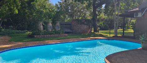 Una piscina azul cristalina rodeada de un patio de ladrillo, con árboles y un muro de ladrillo al fondo.