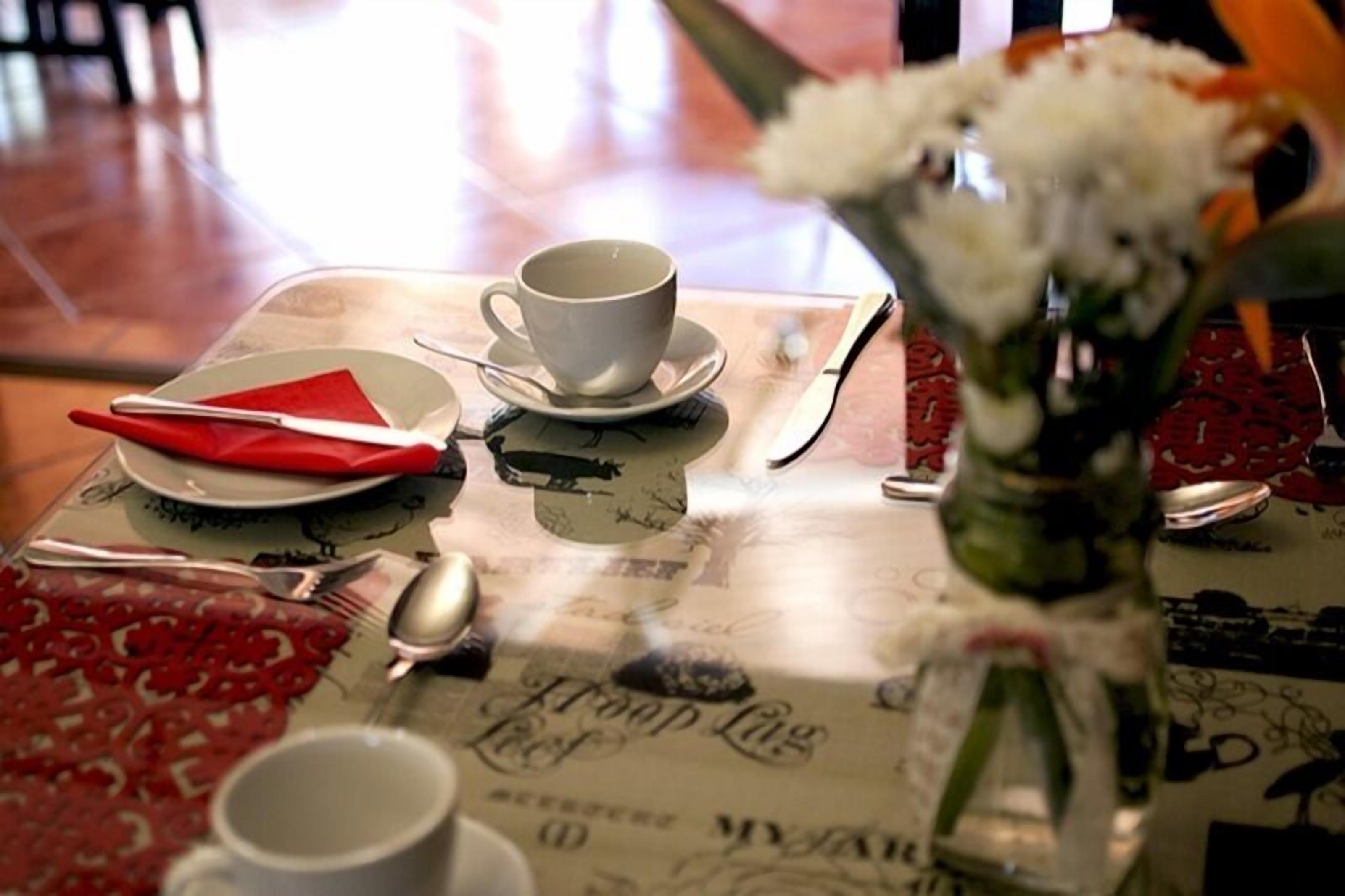 A table set with a white cup, saucer, red napkin, and cutlery. A vase with white flowers is on the table.