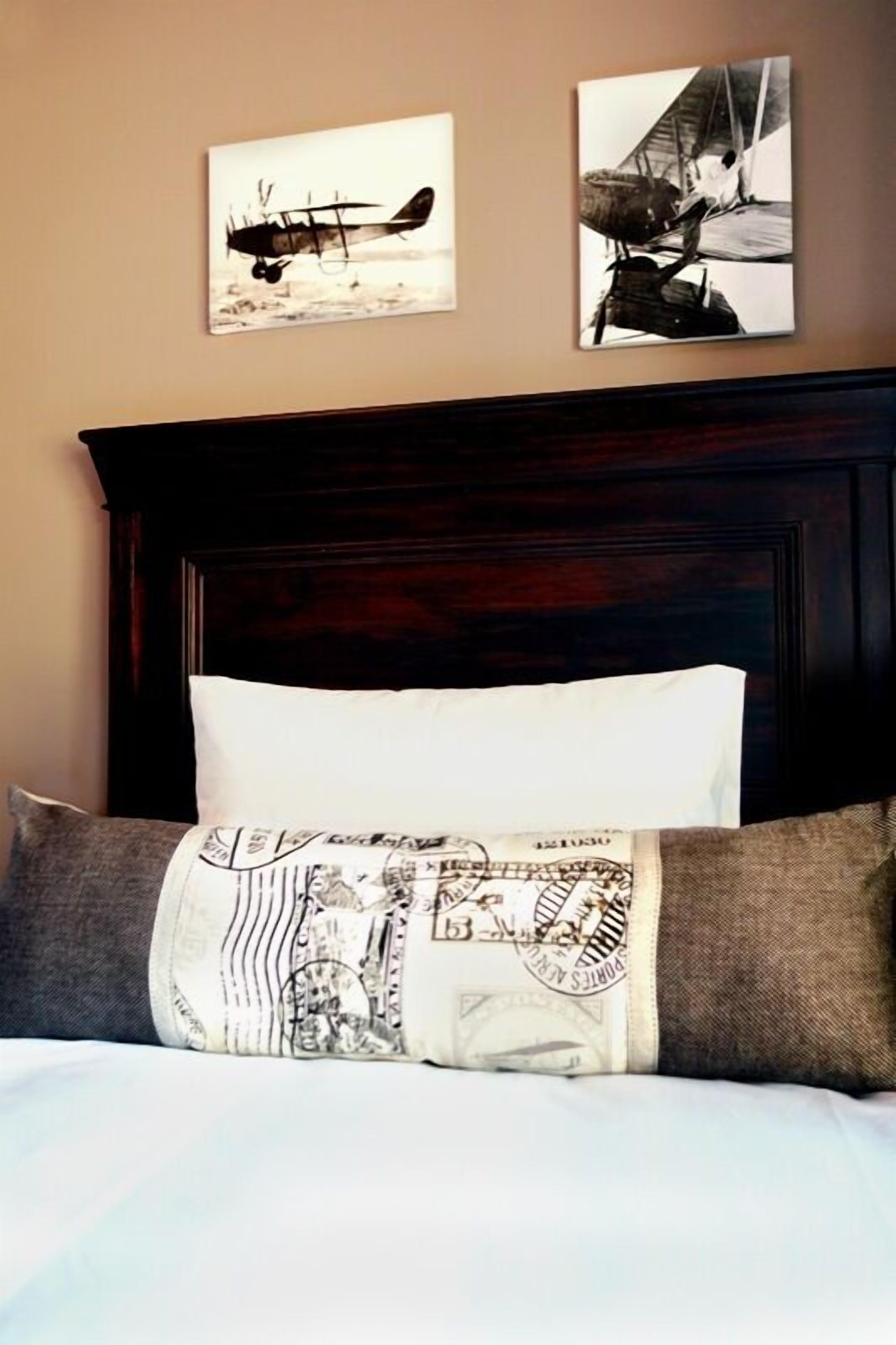Una cama con cabecera de madera oscura, almohada blanca y almohada estampada. Dos cuadros de aviones en blanco y negro adornan la pared sobre la cama.