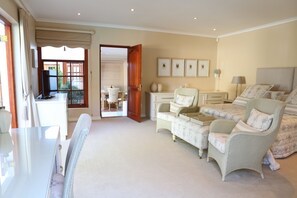 A spacious bedroom with a large bed, a sitting area with a chair and table, and a view through an open door to a balcony.