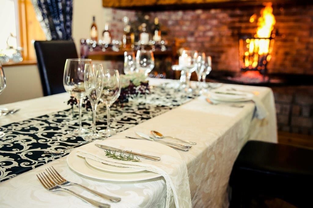 A dining table set with white tablecloth, silverware, and wine glasses, with a patterned runner and a brick wall in the background.