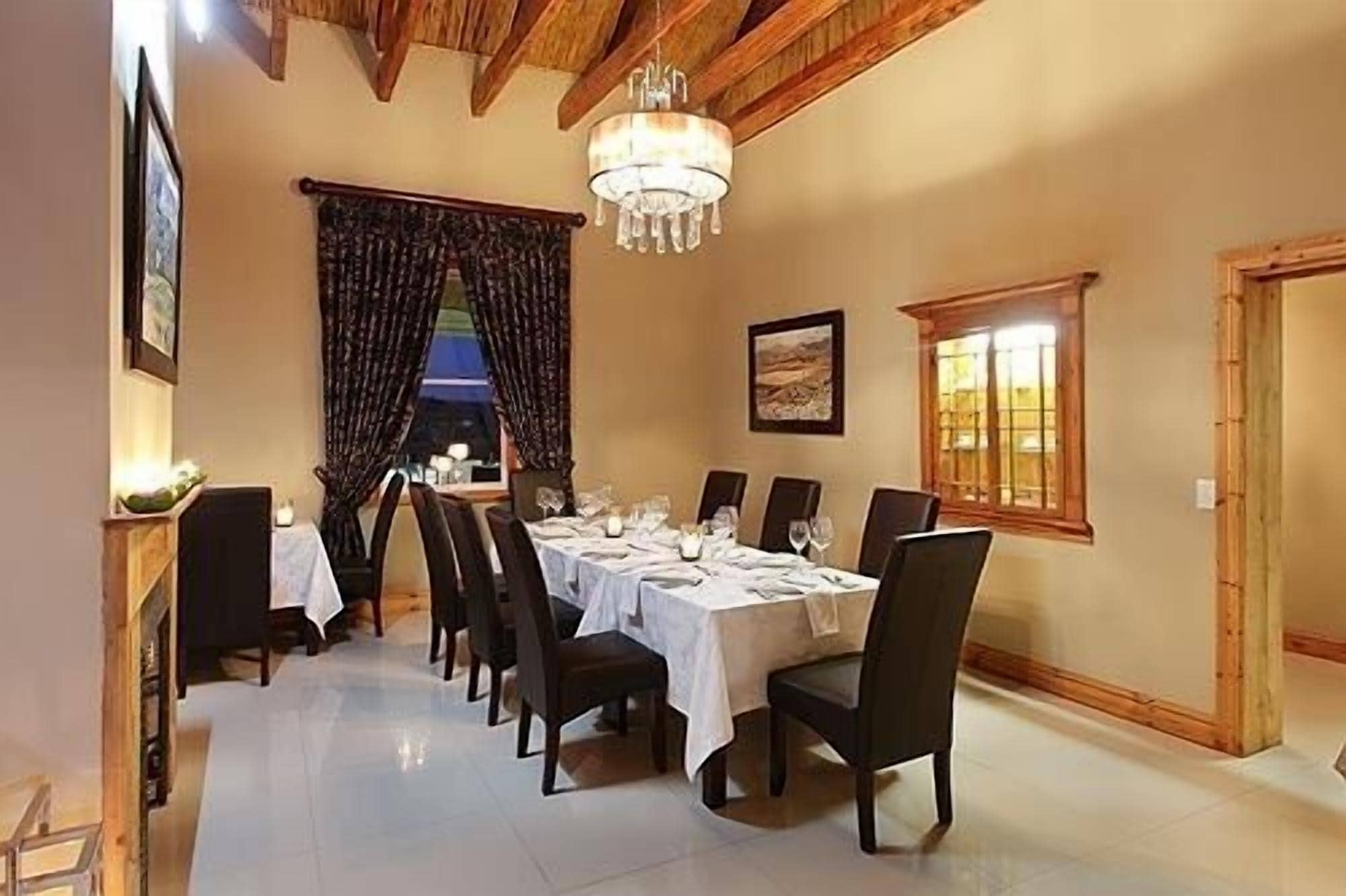 A dining room with a long table set for six, wooden chairs, a chandelier, a window with curtains, and a framed picture on the wall.