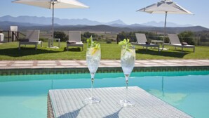 Une scène au bord de la piscine avec deux verres contenant une boisson claire, une table en osier blanche et des chaises longues avec des parasols.