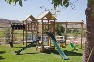 A wooden play structure with a slide and climbing net, surrounded by a white fence.