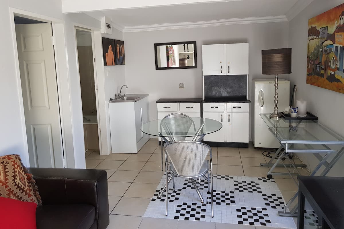 A compact living space with a kitchenette, a dining area with a glass table and metal chairs, and a sofa with a patterned cushion.