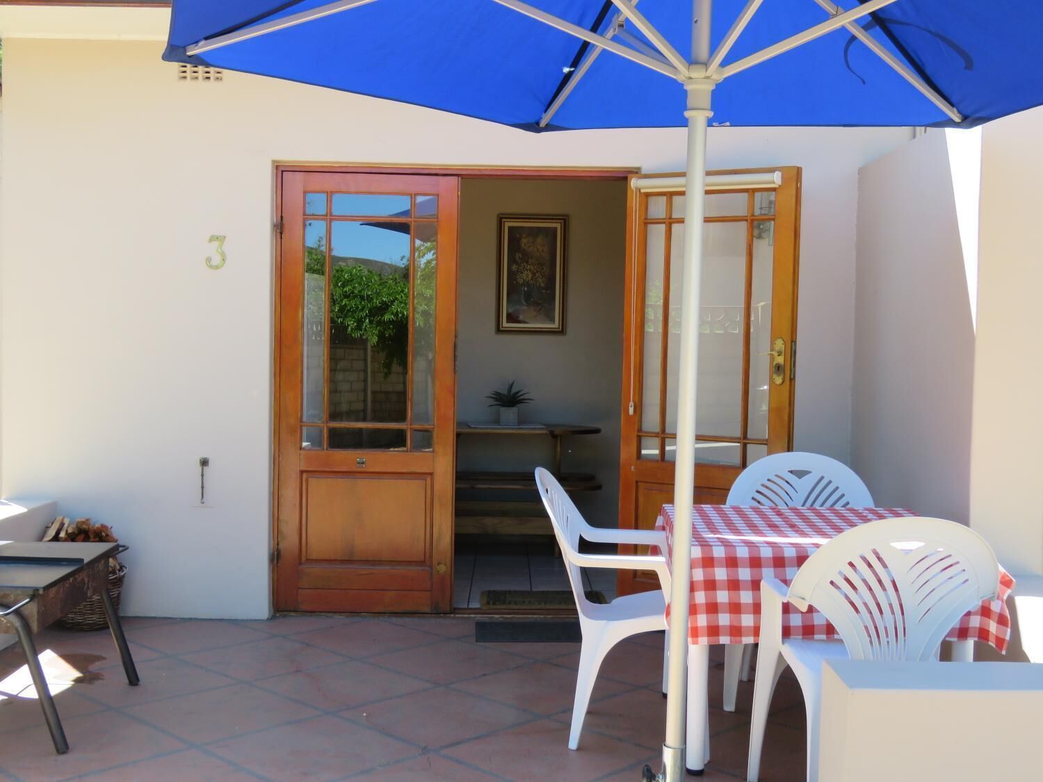 Un patio con mesa y sillas bajo un paraguas azul, una puerta de madera y una pequeña mesa con una planta.