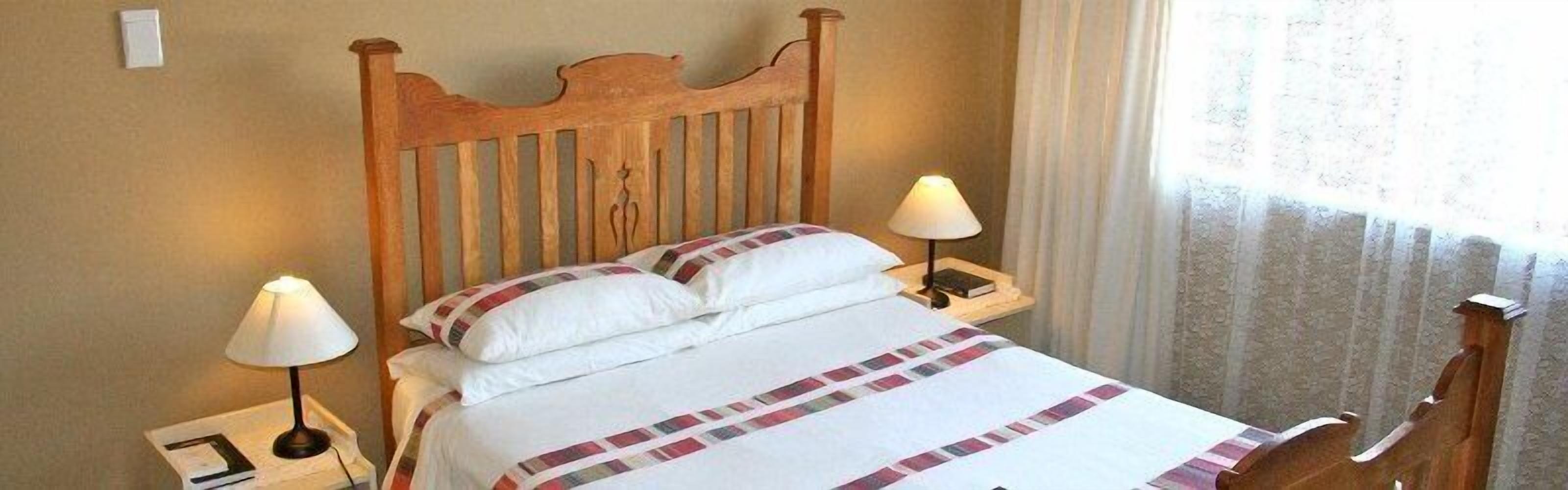 Una cama bien hecha con cabecera de madera, dos lámparas de noche y una ventana con cortinas.