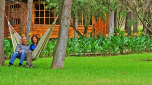 A couple sitting in a hammock in front of a wooden cabin with a garden. - Inraki Lodge (Guayllabamba)